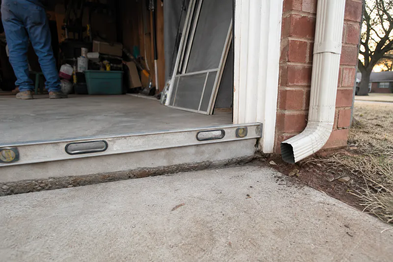 Concrete driveway dropped below the garage slab, with a level spanning the gap and a downspout nearby