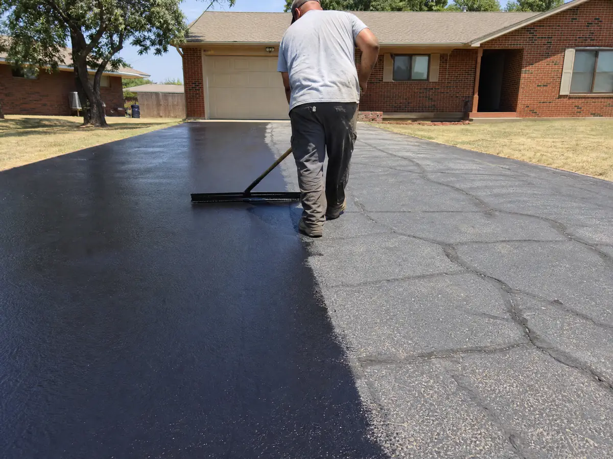 Worker squeegeeing fresh black sealcoat onto an oxidized gray asphalt driveway in Oklahoma summer sun